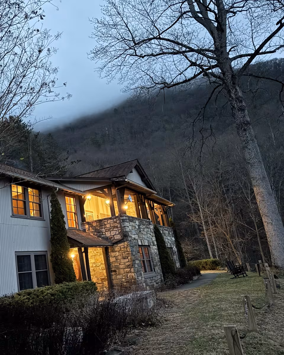 The lodge at dusk with its windows and porch lit and low cloud on the wooded ridge behind